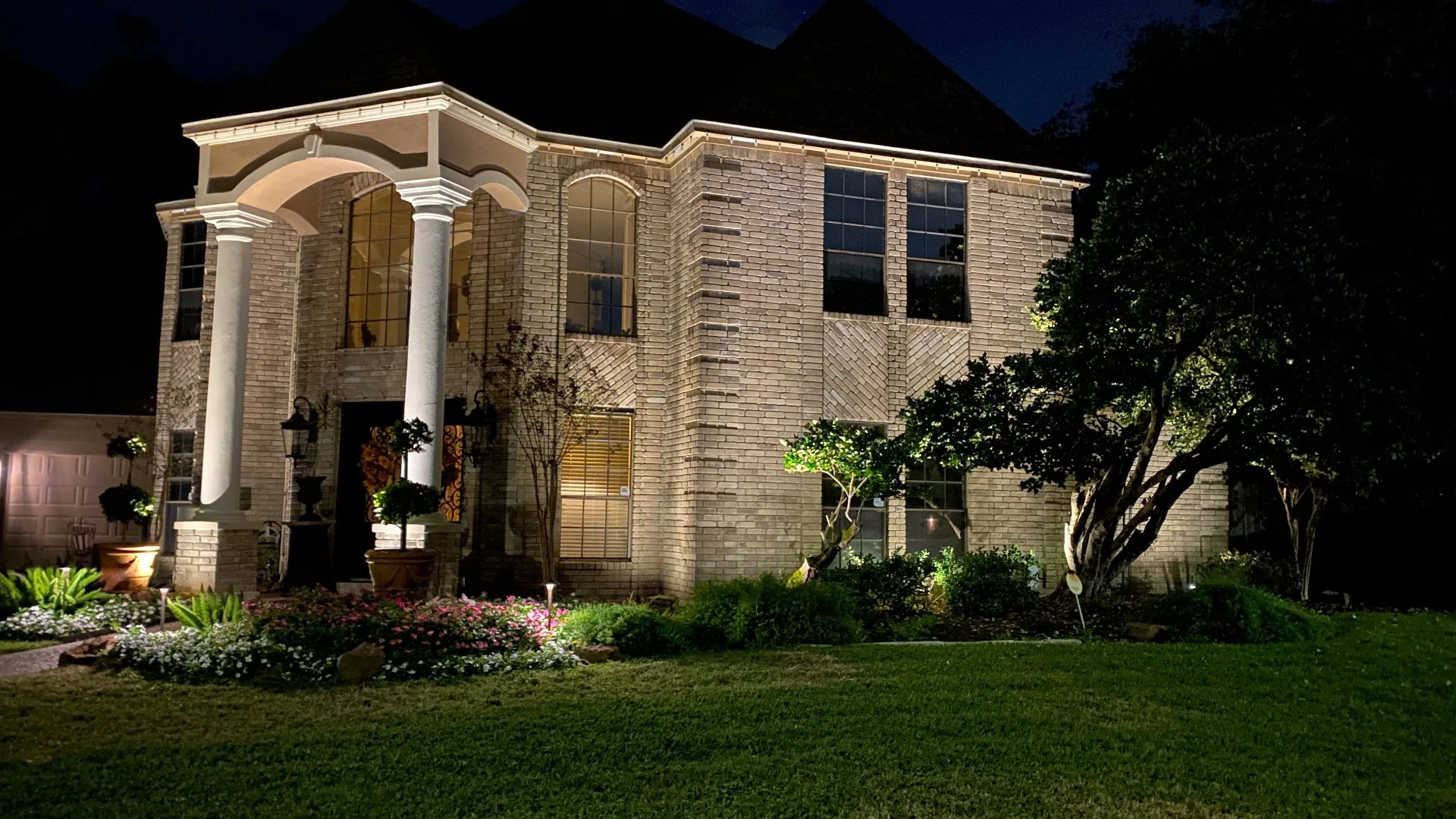 Nighttime view of a well‑lit brick house with tall white columns, large windows, and accent lighting on plants and trees